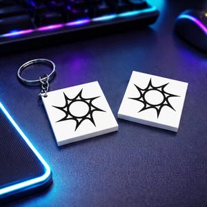 May include: White square keychains and coasters with a black sunburst design. One keychain is attached to a silver ring. The coasters and keychain are on a dark blue surface, with a keyboard and mouse in the background.
