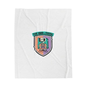 Puede incluir: Manta blanca con un gráfico colorido. El gráfico presenta un diseño de escudo con un esqueleto jugando a un videojuego, una pantalla de ordenador y el texto "THE 1980'S FOSSIL" y "analog original". El escudo tiene acentos naranjas, morados y turquesas.