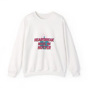 May include: White sweatshirt with the text "HEARTBREAK MADE ME HOTTER" in pink and blue. The text is centered on the front of the sweatshirt. Above the text is a small graphic of a heart and arrow.