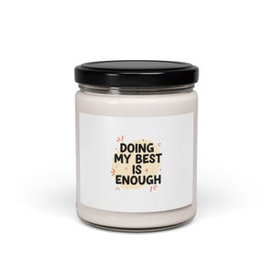 Puede incluir: Una vela blanca en un tarro de cristal con tapa negra. La etiqueta presenta el texto "DOING MY BEST IS ENOUGH" en letras negras y naranjas, con adornos de estrellas.