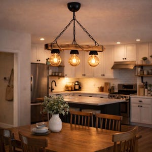 May include: A rustic, three-light chandelier with Edison-style bulbs hangs above a wooden dining table. The fixture features a wooden beam, black chain, and black bulb sockets. The warm light illuminates the kitchen, highlighting the wooden table and white vase.