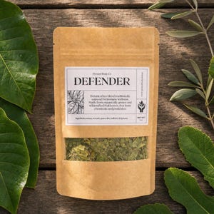 May include: A brown kraft paper pouch containing a botanical tea blend. The label reads DEFENDER, Present Body Co, and lists ingredients including soursop, avocado, guava, olive, mulberry, and fig leaves. The pouch has a clear window showing the dried leaves.