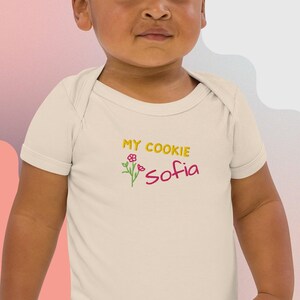 Op de afbeelding: Crèmekleurige babybody met de tekst "MY COOKIE Sofia" in geel en roze. Een klein bloemmotief staat naast de tekst. De body heeft korte mouwen en een eenvoudig, klassiek ontwerp.