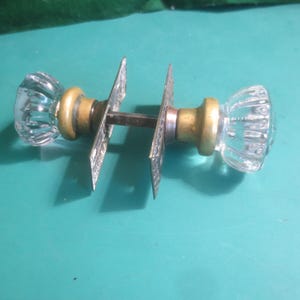 May include: Clear glass door knobs with a ribbed design, brass collars, and metal plates. The hardware is designed for a door and is set against a teal background.