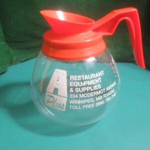 May include: A clear glass coffee carafe with a bright red lid and handle. The carafe has the text "A Plus Restaurant Equipment & Supplies" printed on the side. The carafe is designed for commercial use.