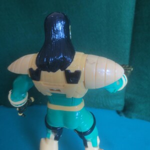 Op de afbeelding: Een plastic actiefiguur van achteren gezien, met lang zwart haar, een beige borstplaat met zwarte accenten en een blauwgroen en beige pak. Het figuur heeft beweegbare schouders en armen, met zichtbare zwarte details op de benen en romp.