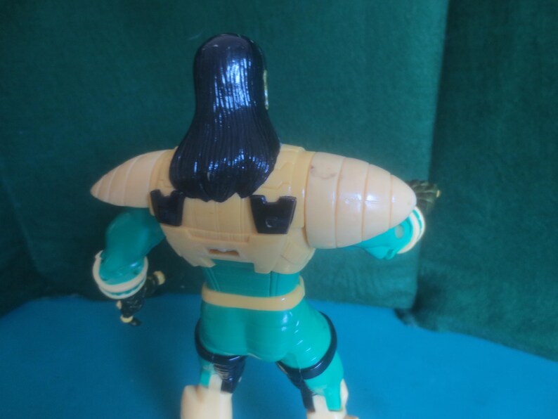 Op de afbeelding: Een plastic actiefiguur van achteren gezien, met lang zwart haar, een beige borstplaat met zwarte accenten en een blauwgroen en beige pak. Het figuur heeft beweegbare schouders en armen, met zichtbare zwarte details op de benen en romp.
