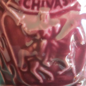 May include: A maroon-colored ceramic container with embossed figures and the word "CHIVAS" at the top. The container has a glossy finish and a textured surface, suggesting it is a decorative item.