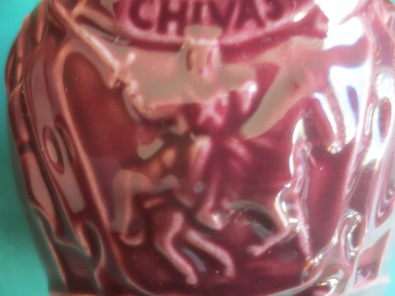 May include: A maroon-colored ceramic container with embossed figures and the word "CHIVAS" at the top. The container has a glossy finish and a textured surface, suggesting it is a decorative item.