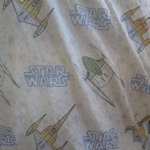 May include: Off-white Star Wars themed fabric featuring repeating patterns of yellow and gray starfighters and the Star Wars logo in light blue. The fabric has a slightly wrinkled texture.