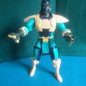 Op de afbeelding: Een plastic actiefiguur van een donkerhuidig personage met lang zwart haar. Het figuur draagt een gele borstplaat, schoudervullingen en laarzen, gecombineerd met een groen pak, zwarte accenten en een gele riemgesp met een W-vormig embleem.