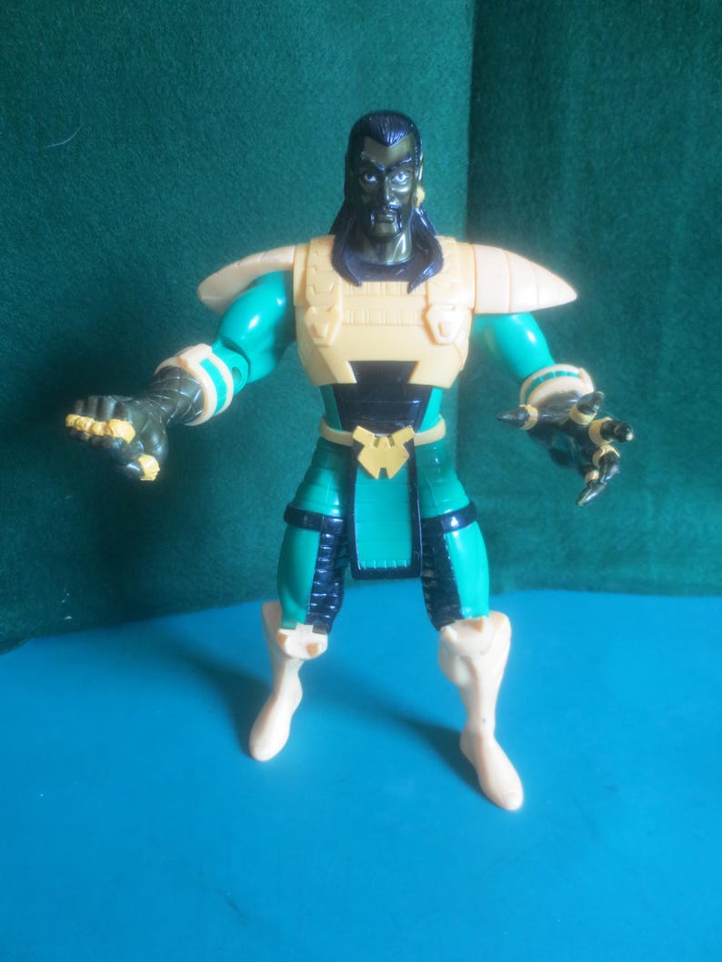 Op de afbeelding: Een plastic actiefiguur van een donkerhuidig personage met lang zwart haar. Het figuur draagt een gele borstplaat, schoudervullingen en laarzen, gecombineerd met een groen pak, zwarte accenten en een gele riemgesp met een W-vormig embleem.