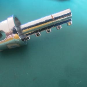 Puede incluir: Primer plano de una llave plateada y brillante. La llave tiene un bucle circular en un extremo y una serie de muescas rectangulares a lo largo de su borde. La llave est&aacute; sobre una superficie de color turquesa.