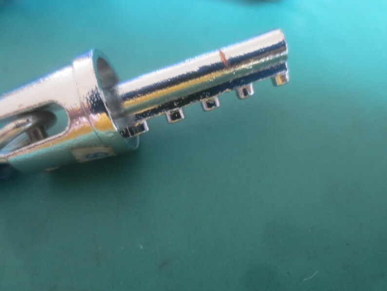 Puede incluir: Primer plano de una llave plateada y brillante. La llave tiene un bucle circular en un extremo y una serie de muescas rectangulares a lo largo de su borde. La llave est&aacute; sobre una superficie de color turquesa.
