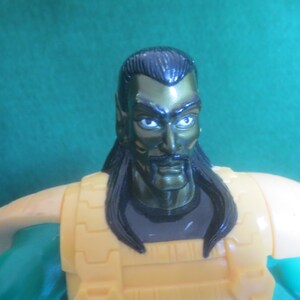 Op de afbeelding: Een plastic actiefiguur met een donkergroen metallic hoofd, puntige oren, zwart haar en een snor. Het figuur draagt een gele borstplaat met schoudervullingen over een donkergrijs shirt en heeft groene mouwen, gefotografeerd vanaf de borst.