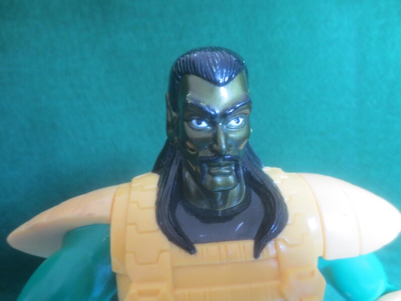 Op de afbeelding: Een plastic actiefiguur met een donkergroen metallic hoofd, puntige oren, zwart haar en een snor. Het figuur draagt een gele borstplaat met schoudervullingen over een donkergrijs shirt en heeft groene mouwen, gefotografeerd vanaf de borst.
