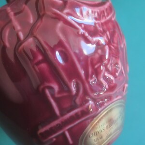 May include: A close-up of a burgundy-colored ceramic bottle with embossed detailing. The bottle has a rounded shape and a glossy finish. A gold label with text is visible. The bottle is likely for spirits or liquor.