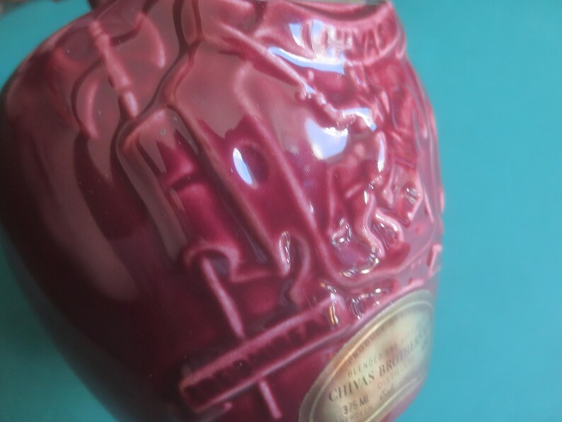 May include: A close-up of a burgundy-colored ceramic bottle with embossed detailing. The bottle has a rounded shape and a glossy finish. A gold label with text is visible. The bottle is likely for spirits or liquor.