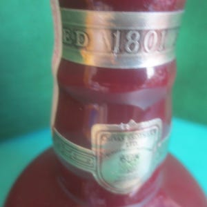 May include: Close-up of a burgundy-colored bottle with silver accents. The bottle has a silver band with the text "ED 1801". A silver label with text is also visible. The background is a mix of green and blue.