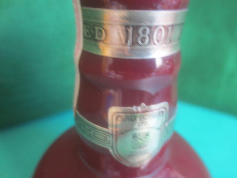 May include: Close-up of a burgundy-colored bottle with silver accents. The bottle has a silver band with the text "ED 1801". A silver label with text is also visible. The background is a mix of green and blue.