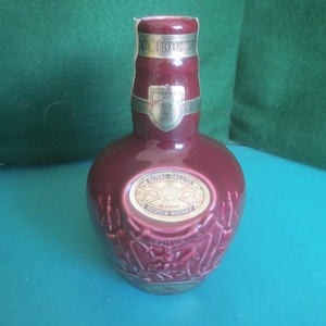 May include: A maroon-colored Royal Salute Scotch Whisky bottle. The bottle has a gold label with the brand name and a decorative embossed design. The bottle is on a blue surface.