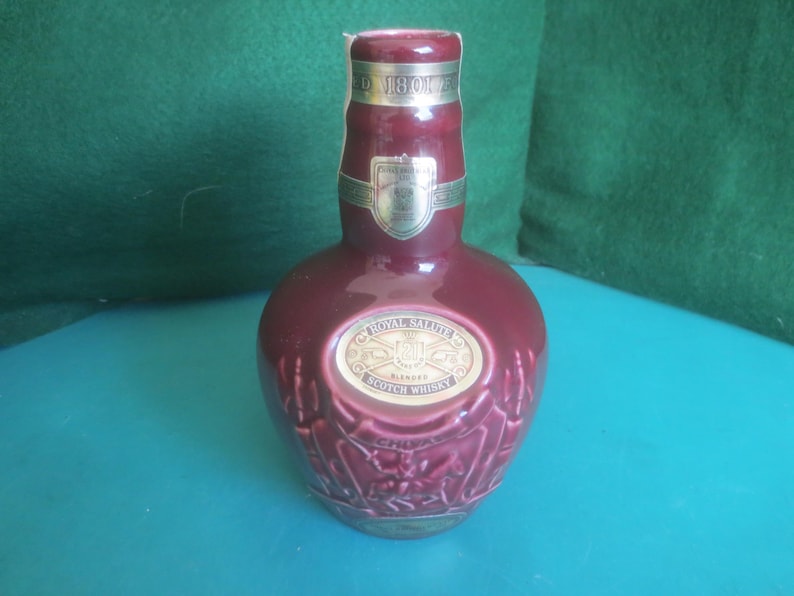 May include: A maroon-colored Royal Salute Scotch Whisky bottle. The bottle has a gold label with the brand name and a decorative embossed design. The bottle is on a blue surface.