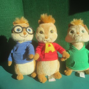 May include: Three plush chipmunk toys. One wears a blue hoodie and glasses, the second a red hoodie with a yellow "A", and the third a green hoodie. All have tan bodies and brown hair.