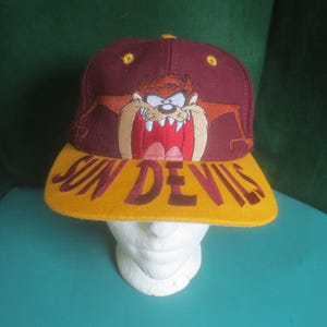 May include: A maroon baseball cap featuring an embroidered graphic of the Tasmanian Devil character on the front panel. The flat brim is yellow and displays the text SUN DEVILS in maroon lettering. The cap includes yellow eyelet vents.