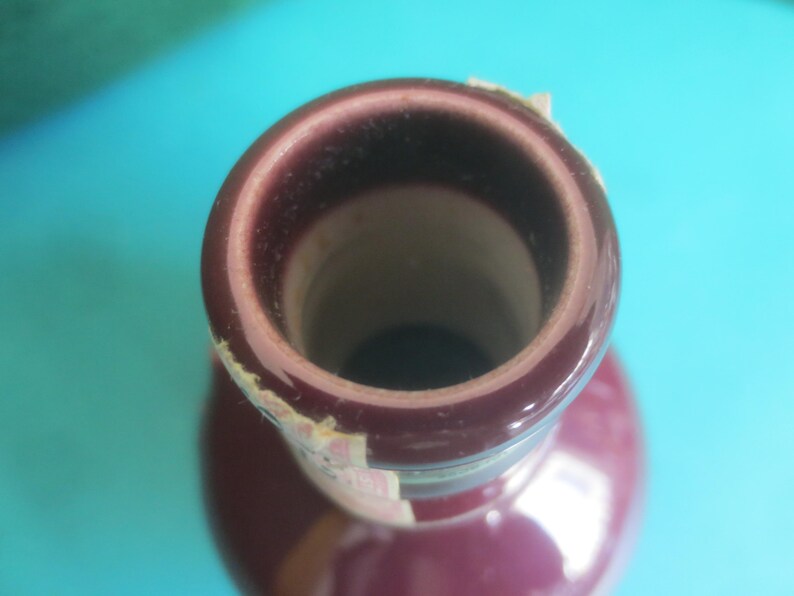 May include: Close-up of a maroon-colored bottle neck with a dark interior. The rim of the bottle has a lighter, reddish-brown hue. A partially visible label is affixed to the side of the bottle.