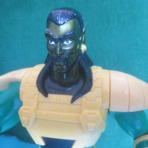 Op de afbeelding: Een plastic actiefiguur met een donkergroen gezicht, zwart haar, een snor en puntige oren. Het figuur draagt een gele borstplaat met gestructureerde details en bijpassende gele schouderstukken over groene armen. De uitdrukking is serieus.