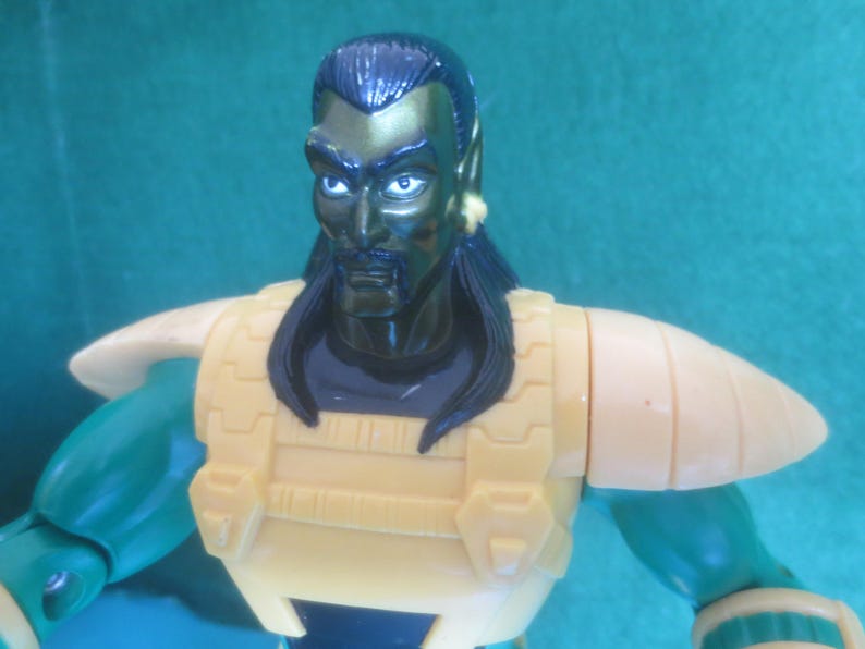 Op de afbeelding: Een plastic actiefiguur met een donkergroen gezicht, zwart haar, een snor en puntige oren. Het figuur draagt een gele borstplaat met gestructureerde details en bijpassende gele schouderstukken over groene armen. De uitdrukking is serieus.
