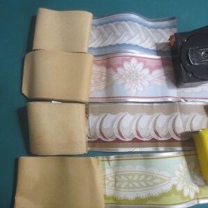 May include: An assortment of wallpaper border rolls in various patterns and colors, including floral and wavy designs. A black tape measure and a yellow utility knife are also present, suggesting a home improvement or crafting theme.