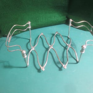 May include: A silver-colored, wire kitchen utensil with a zig-zag design. The utensil has two curved handles and is designed to hold something. The object is on a teal surface with a green background.