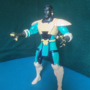 Op de afbeelding: Een plastic actiefiguur met een donkere huid, een sikje en lang zwart haar. Het figuur draagt een groenblauw en zwart pak met beige pantserplaten op de schouders, borst, taille en schenen, en heeft klauwvormige handen in een actieve houding.