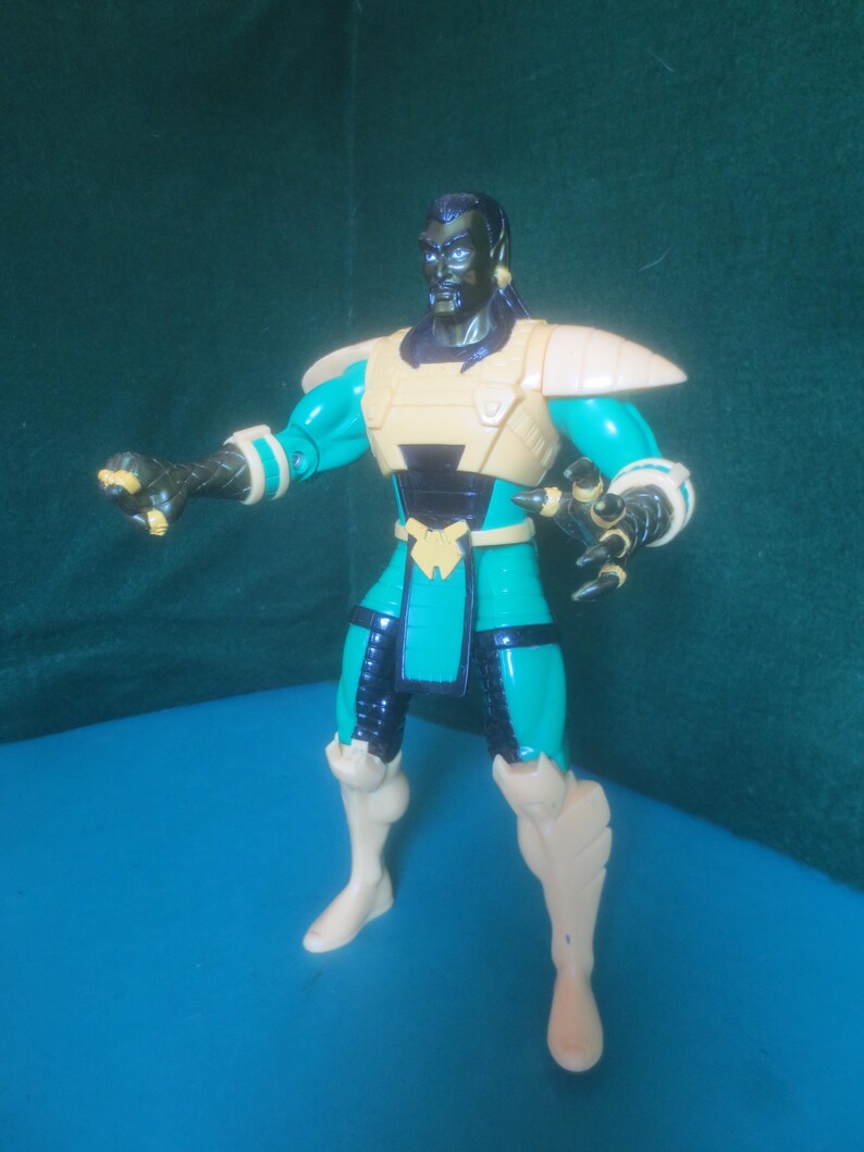 Op de afbeelding: Een plastic actiefiguur met een donkere huid, een sikje en lang zwart haar. Het figuur draagt een groenblauw en zwart pak met beige pantserplaten op de schouders, borst, taille en schenen, en heeft klauwvormige handen in een actieve houding.