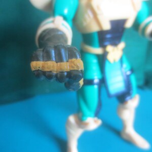 Op de afbeelding: Een plastic actiefiguur in een blauwgroen en cr&egrave;mekleurig pak met zwarte accenten. Het figuur heeft een gebalde vuist naar voren gestoken, met beige details op de knokkels. De romp is voorzien van een donkere borstplaat en een riem, met cr&egrave;mekleurige laarzen en armbanden zichtbaar op de ledematen.