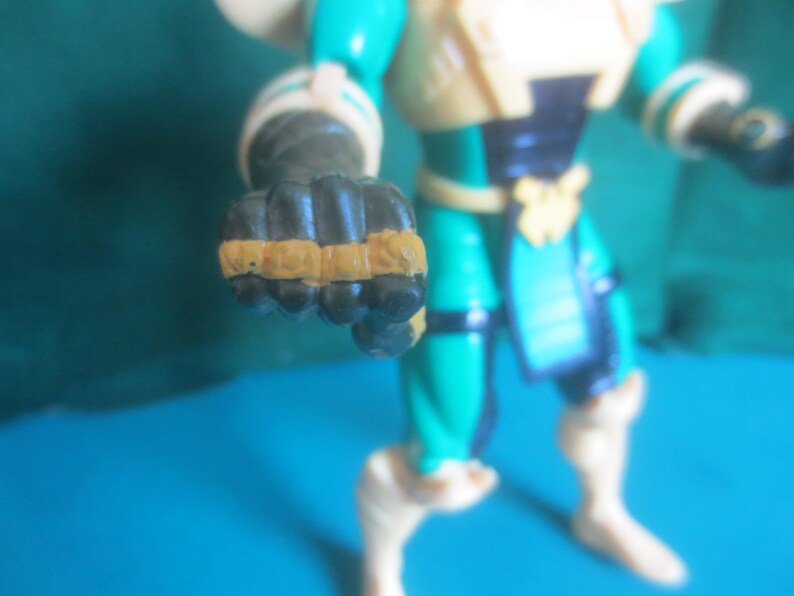 Op de afbeelding: Een plastic actiefiguur in een blauwgroen en cr&egrave;mekleurig pak met zwarte accenten. Het figuur heeft een gebalde vuist naar voren gestoken, met beige details op de knokkels. De romp is voorzien van een donkere borstplaat en een riem, met cr&egrave;mekleurige laarzen en armbanden zichtbaar op de ledematen.