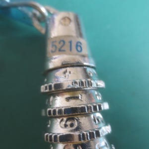 Puede incluir: Primer plano de un candado plateado con el n&uacute;mero "5216" en azul. El candado tiene m&uacute;ltiples diales giratorios con n&uacute;meros y marcas. El fondo es de color verde azulado s&oacute;lido.