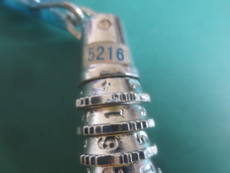 Puede incluir: Primer plano de un candado plateado con el n&uacute;mero "5216" en azul. El candado tiene m&uacute;ltiples diales giratorios con n&uacute;meros y marcas. El fondo es de color verde azulado s&oacute;lido.