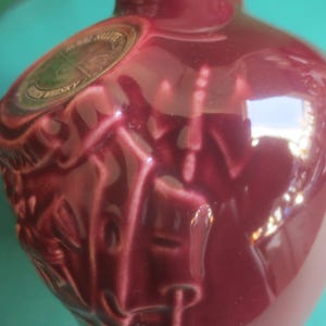 May include: A burgundy-colored ceramic bottle with a glossy finish. The bottle has embossed details on the front and a circular label with gold and green accents. The bottle's shape is rounded, with a narrow neck.