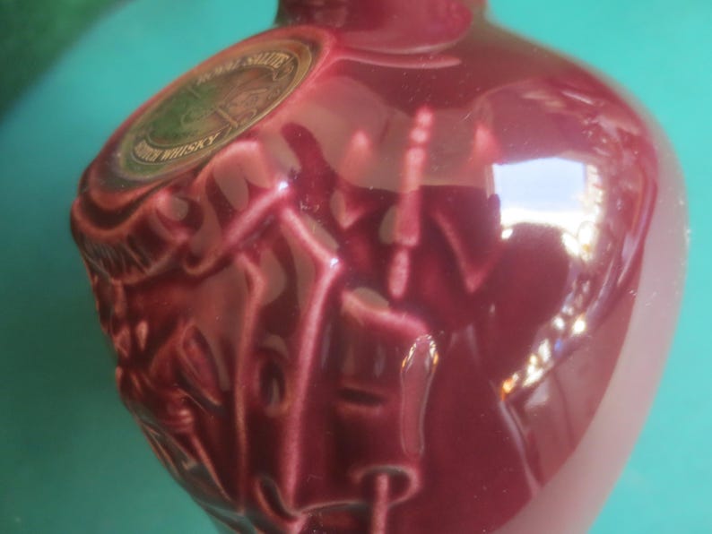 May include: A burgundy-colored ceramic bottle with a glossy finish. The bottle has embossed details on the front and a circular label with gold and green accents. The bottle's shape is rounded, with a narrow neck.