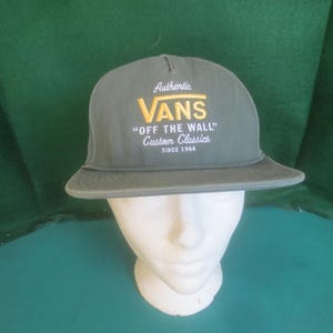 May include: A green flat-brimmed cap with the Vans logo in yellow and white. The cap features the text "Authentic VANS OFF THE WALL Custom Classics SINCE 1966". The cap is displayed on a white mannequin head.