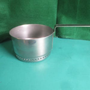 May include: A small, stainless steel saucepan with a long, straight handle. The pot has a rounded body with a series of small holes near the base. The handle extends horizontally from the side of the pot.