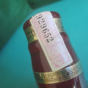 May include: Close-up of a burgundy-colored bottle with a gold band and a pink and white label. The label has the number "929652" printed on it. The bottle is partially visible against a teal background.