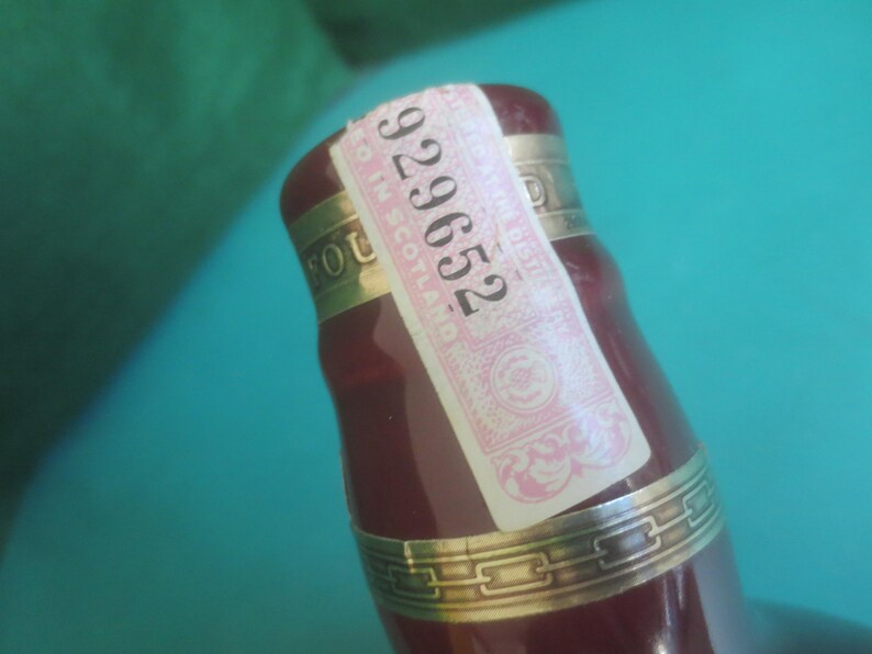 May include: Close-up of a burgundy-colored bottle with a gold band and a pink and white label. The label has the number "929652" printed on it. The bottle is partially visible against a teal background.