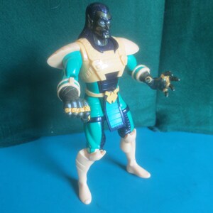 Op de afbeelding: Een plastic actiefiguur met een donkerhuidig personage met lang zwart haar, puntige oren en blauwe ogen. Het figuur draagt een beige borstplaat, een blauwgroen en zwart pak, beige schouderbeschermers en beige laarzen met donkere handschoenen aan de handen.