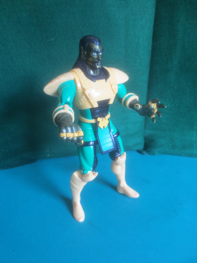 Op de afbeelding: Een plastic actiefiguur met een donkerhuidig personage met lang zwart haar, puntige oren en blauwe ogen. Het figuur draagt een beige borstplaat, een blauwgroen en zwart pak, beige schouderbeschermers en beige laarzen met donkere handschoenen aan de handen.