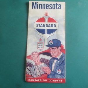 Könnte beinhalten: Vintage Minnesota Reisebroschüre der Standard Oil Company. Die Broschüre zeigt ein rot-weiß-blaues Standard Oil-Logo mit einer Fackel. Das Cover zeigt einen Mann und eine Frau, die sich eine Karte ansehen. Der Text "As You Travel Ask Us" ist ebenfalls sichtbar.