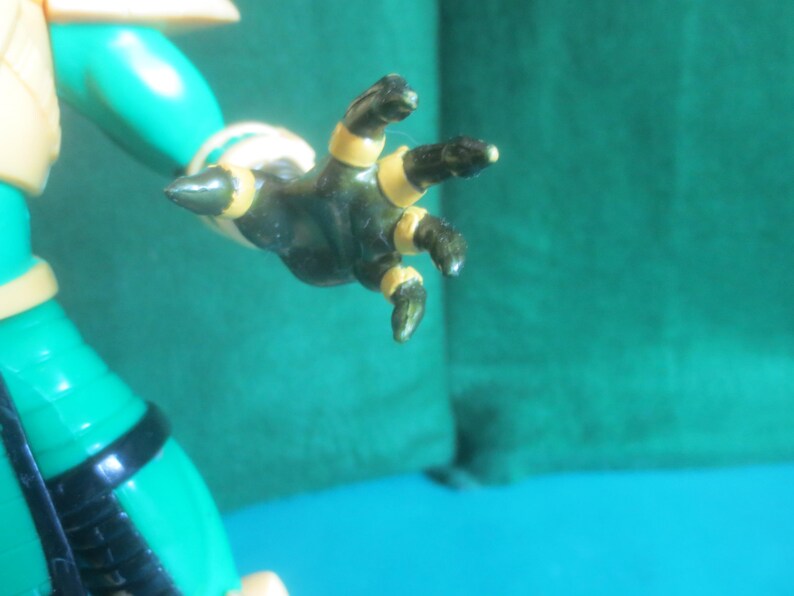Op de afbeelding: Een groene actiefiguurhand met donkergroene vingers en puntige uiteinden, voorzien van gouden bandjes rond elke vinger en de pols. De hand is uitgestrekt en bevestigd aan een groene arm met een zwarte band en een gouden schouderstuk.