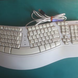 May include: Off-white ergonomic Microsoft keyboard with a curved design. The keyboard has a built-in wrist rest and a standard QWERTY layout. A white cable with a USB connector is attached.
